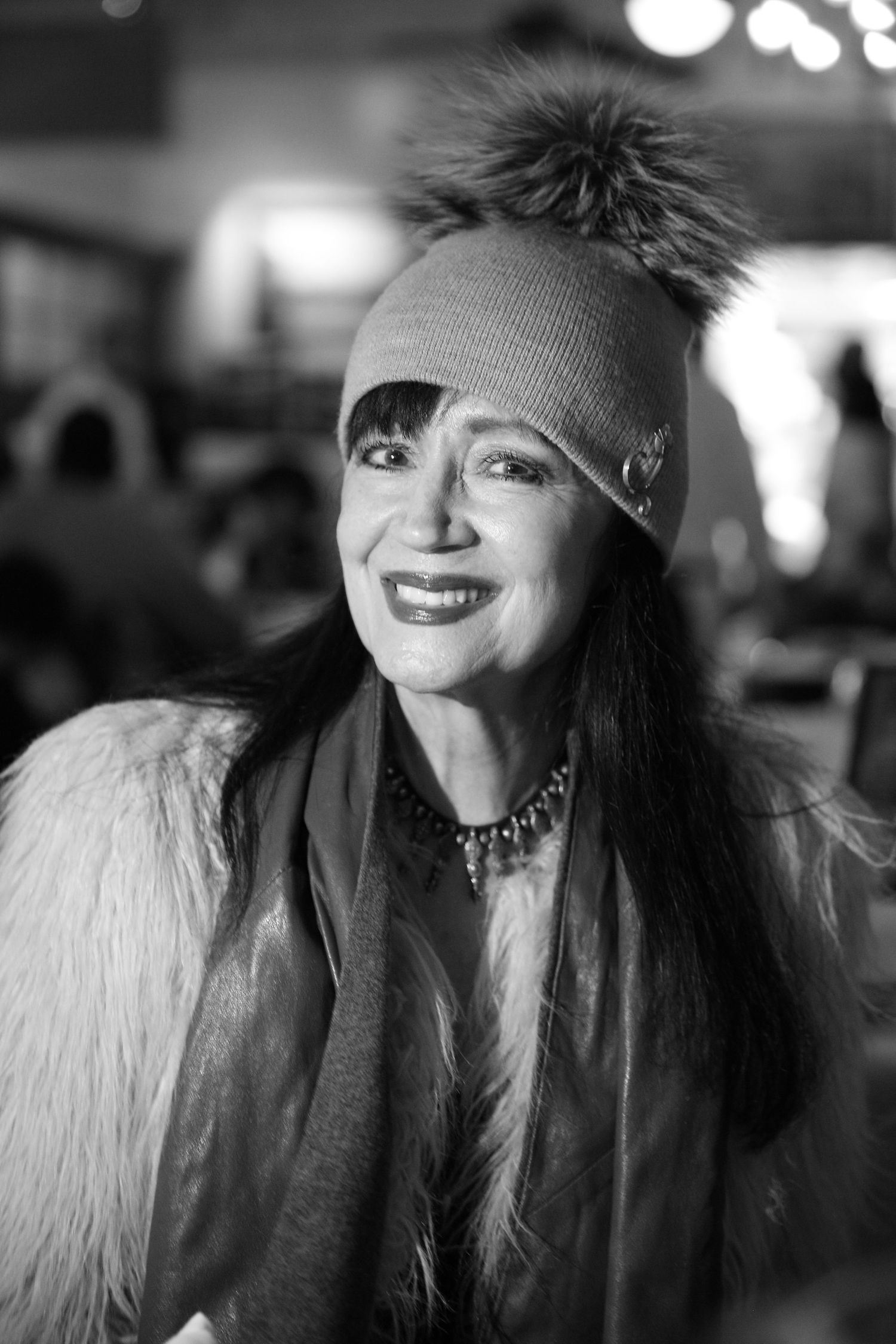 A black and white portrait of someone wearing a knit beanie and furry white coat while smiling at night.