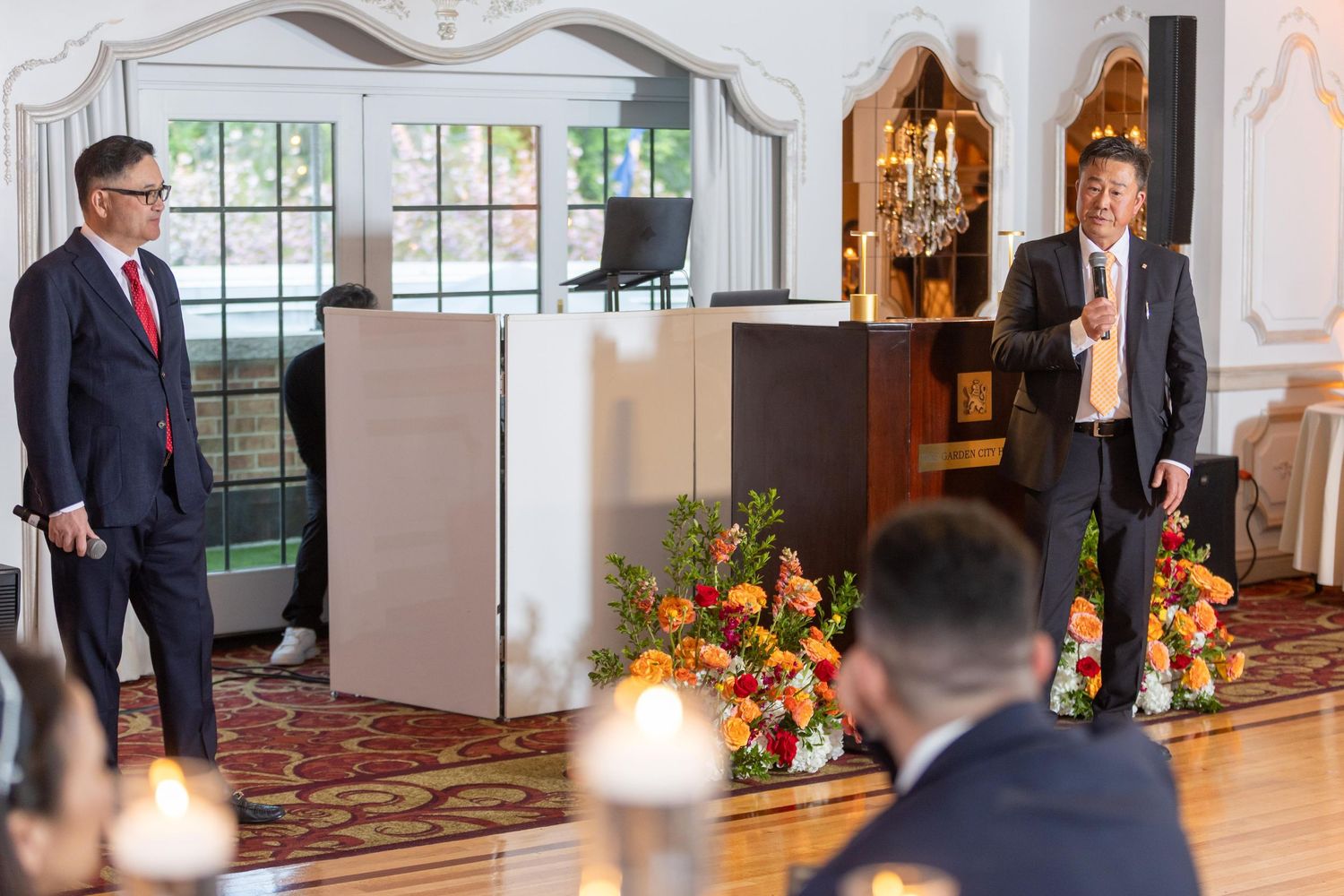 Two men in suits stand near floral arrangements in an elegant room during a formal event.
