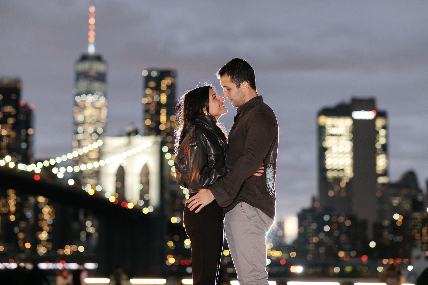 Couple sharing a romantic moment with NYC skyline and Brooklyn Bridge illuminated at night in the background.