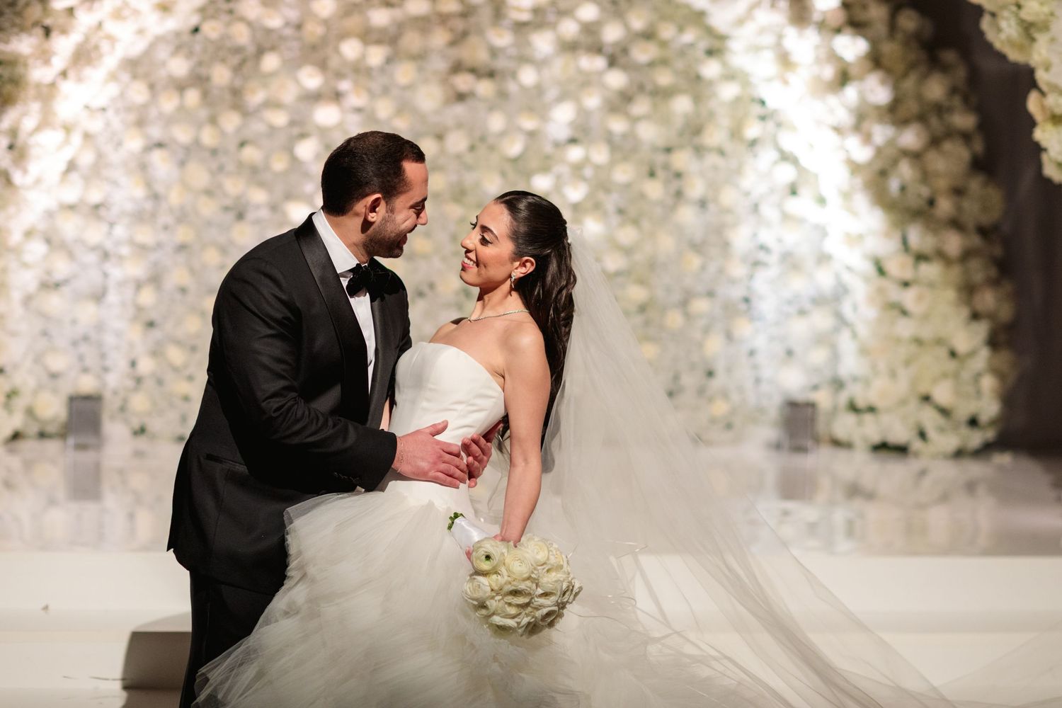 Bride and groom share a romantic moment at elegant wedding ceremony with white floral backdrop.