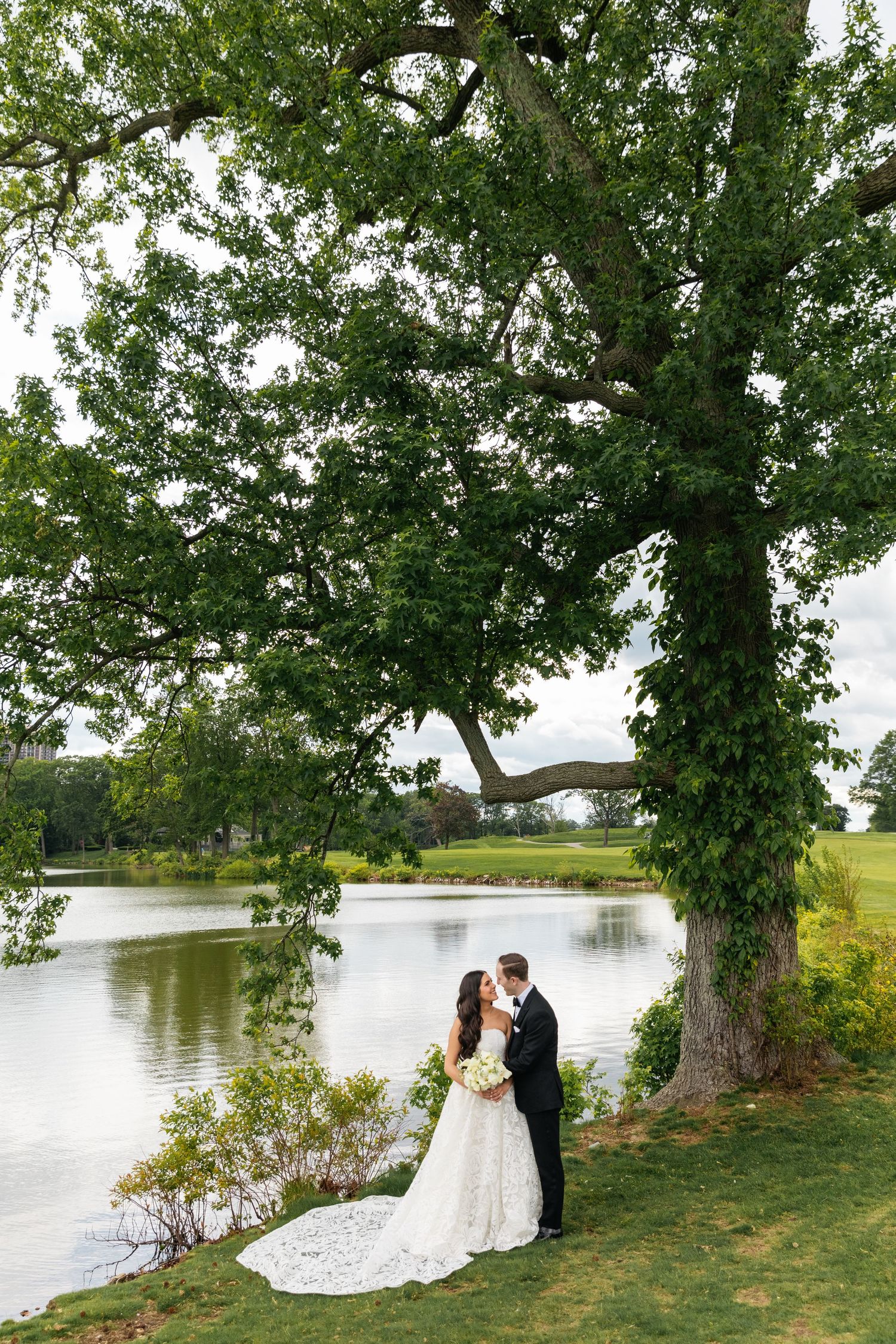 Bride and groom share a romantic moment under a large oak tree beside a serene lake on their wedding day.