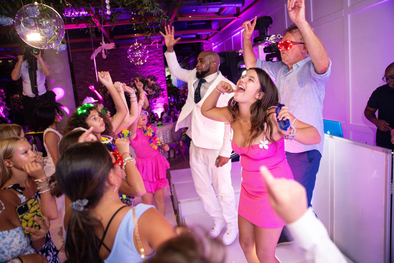 Energetic dance party with guests in colorful outfits, pink lighting, and a performer in white suit holding microphone.