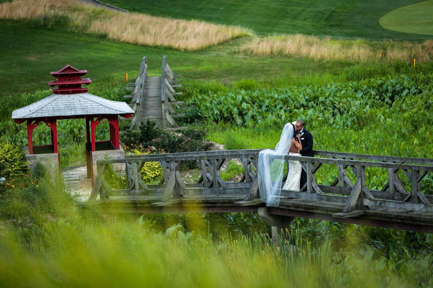 Wedding couple shares romantic moment on ornate bridge with red gazebo in background.