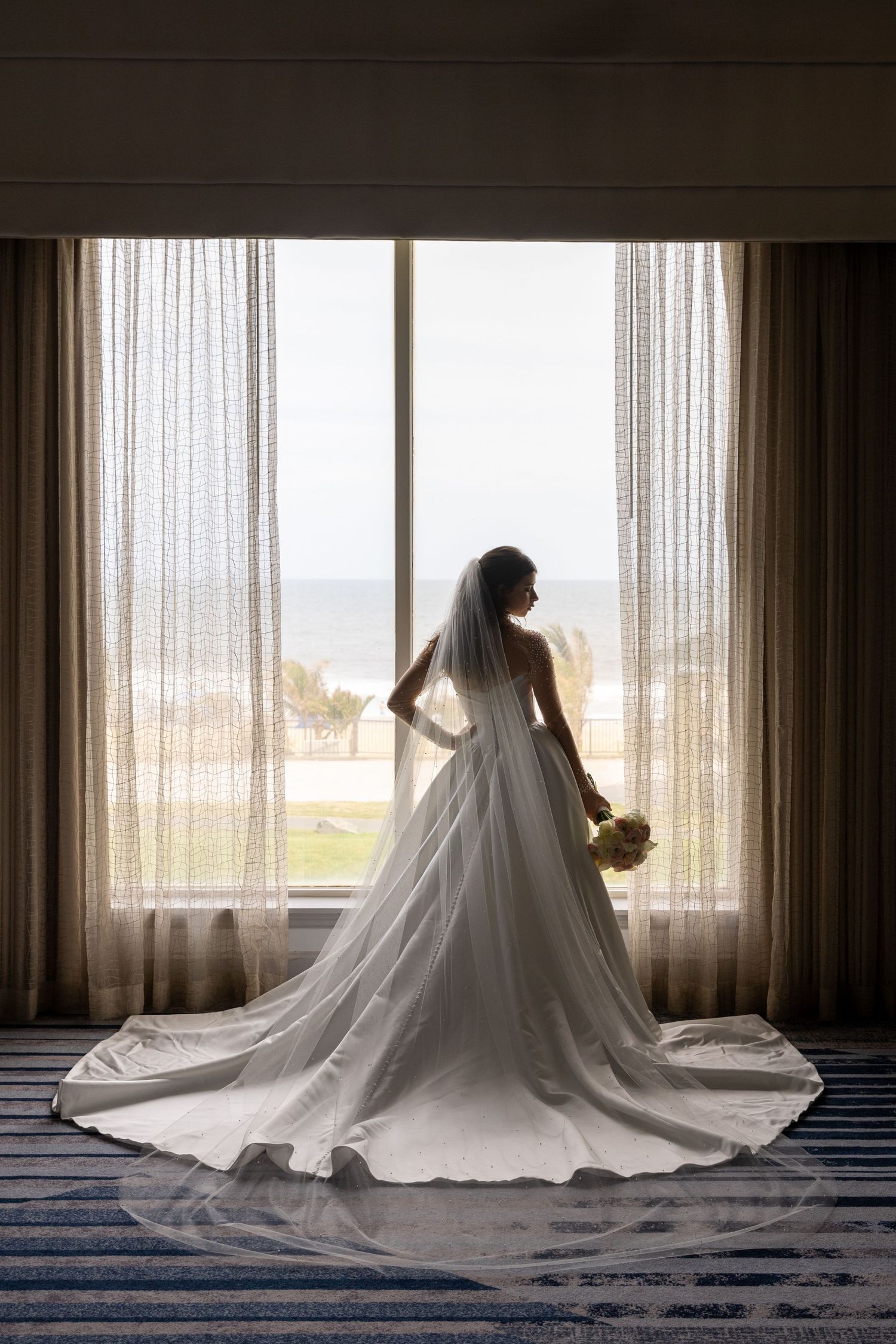 A bride in a flowing white gown with long train stands silhouetted against a window overlooking the ocean.