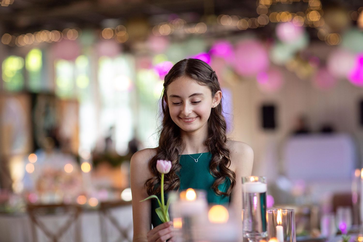 Young woman in teal dress smiling while holding a pink tulip at an elegant candlelit event venue.