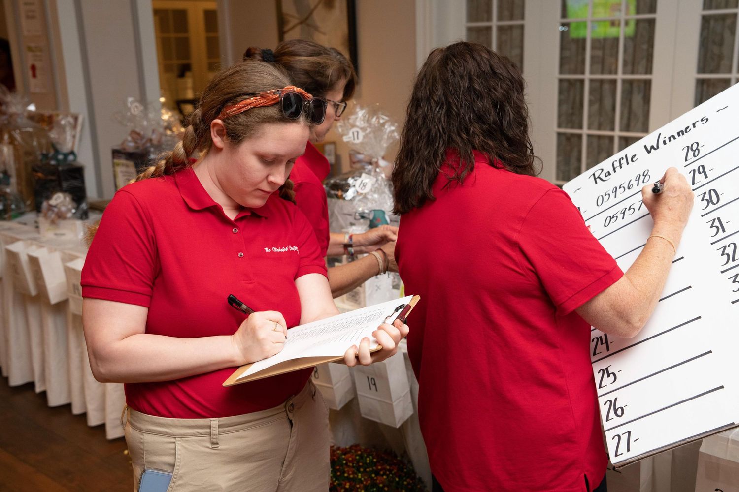 Two women in red polo shirts record raffle winners on a whiteboard at an indoor fundraising event.
