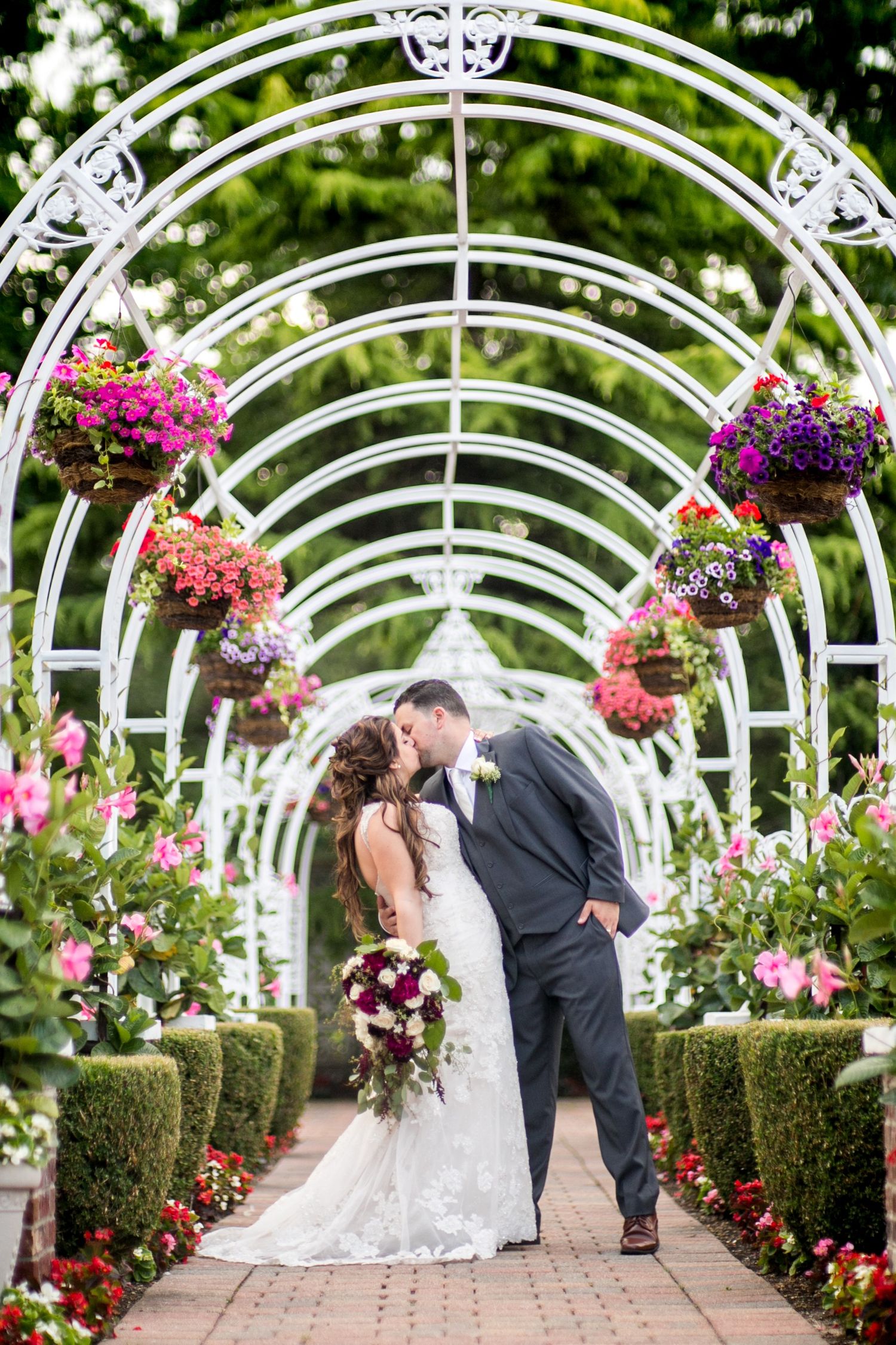 Bride and groom kissing under a white floral arch with hanging flower baskets in a lush garden setting.