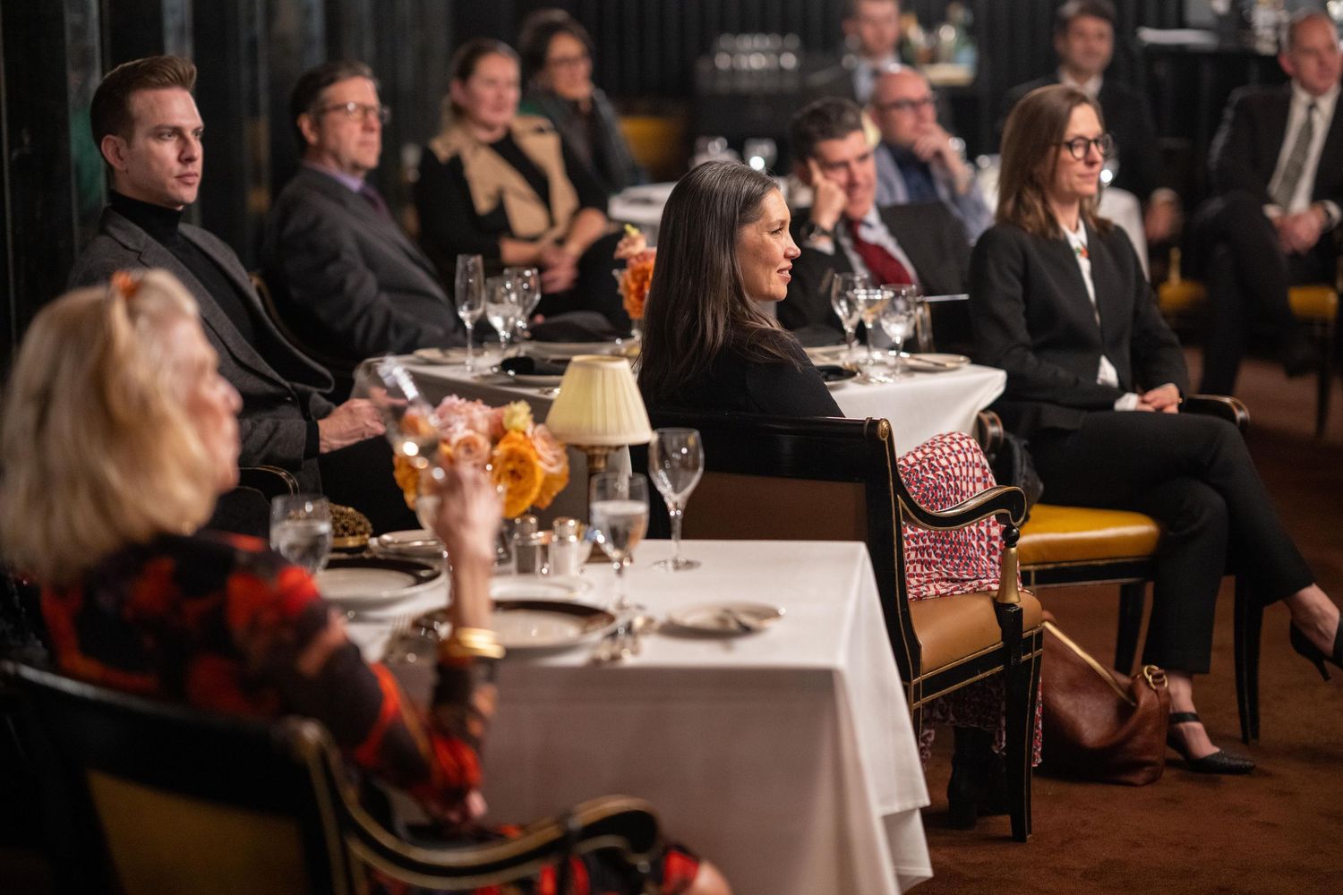 Elegantly dressed guests seated at formal dinner tables during an upscale corporate event in a dimly lit restaurant.