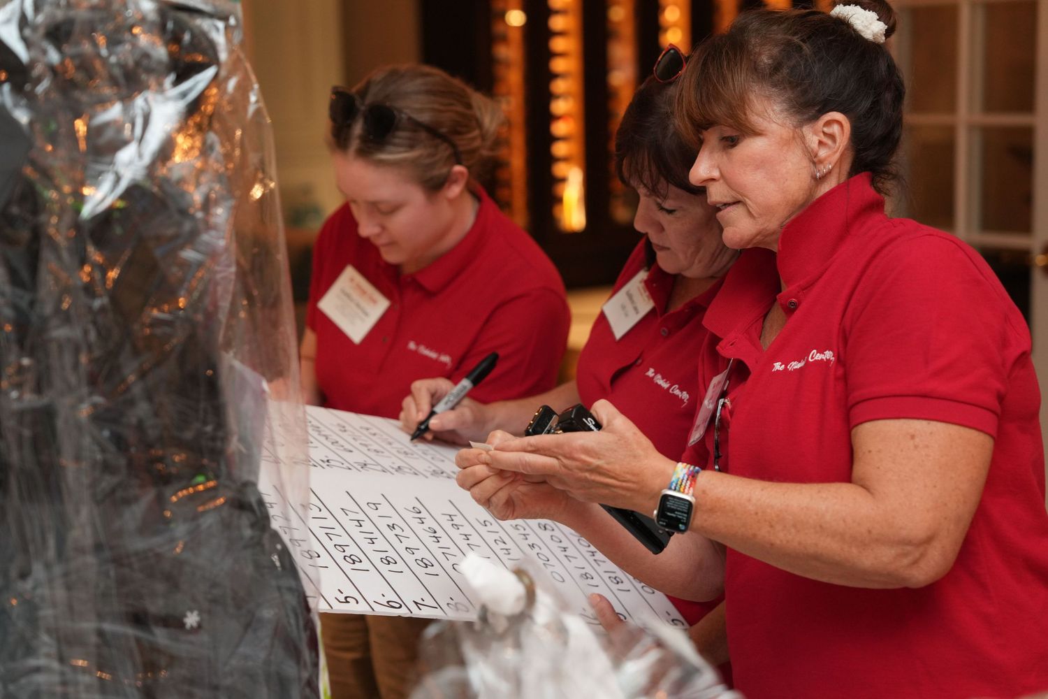 Three women in red polo shirts reviewing and signing documents at a registration table during an event.