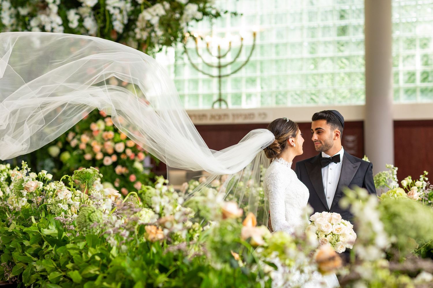 Wedding couple shares romantic moment under flowing veil with menorah and floral decor.
