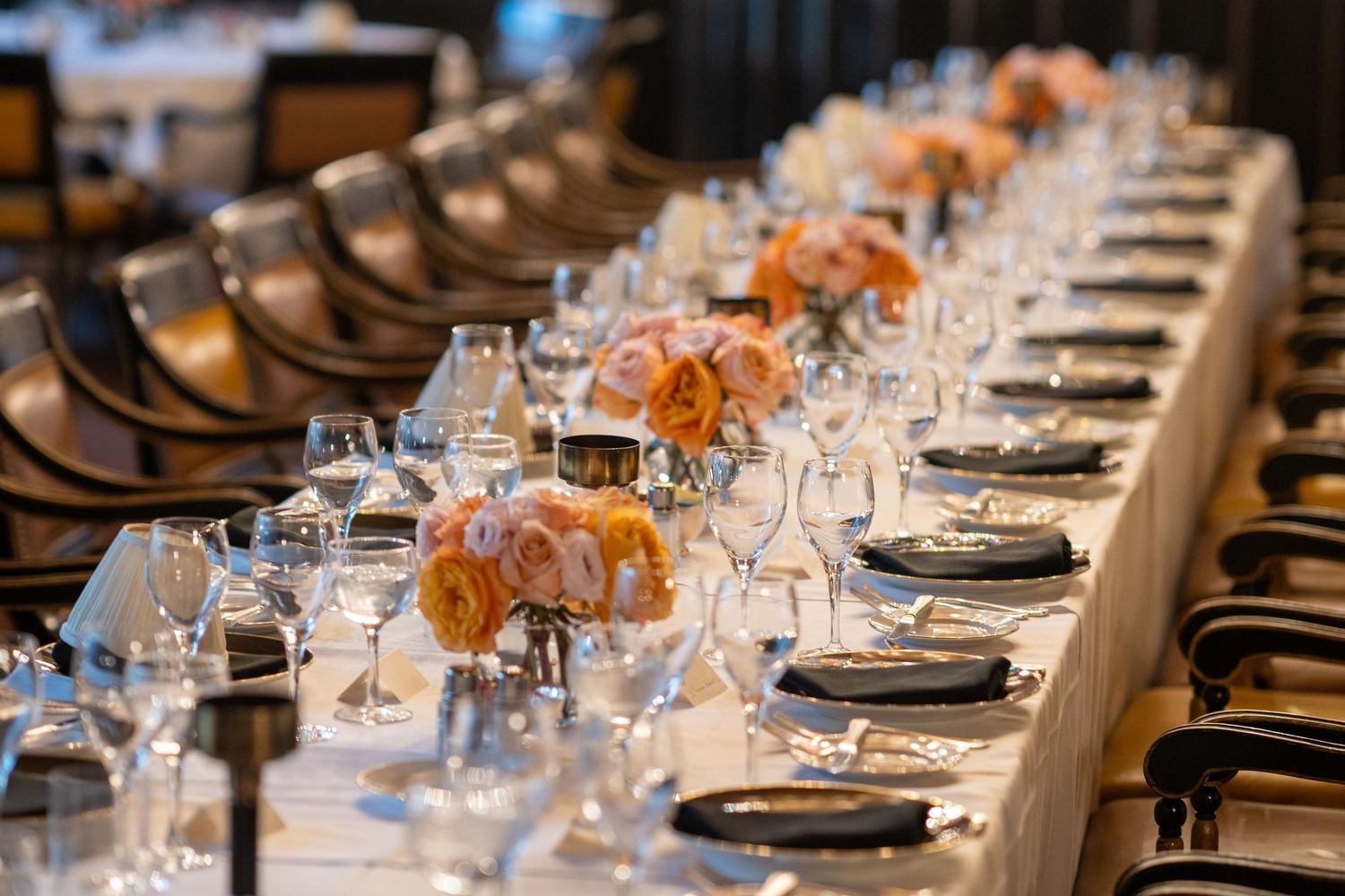 Elegant long banquet table set with black plates, crystal glasses, and peach rose centerpieces for a formal event.