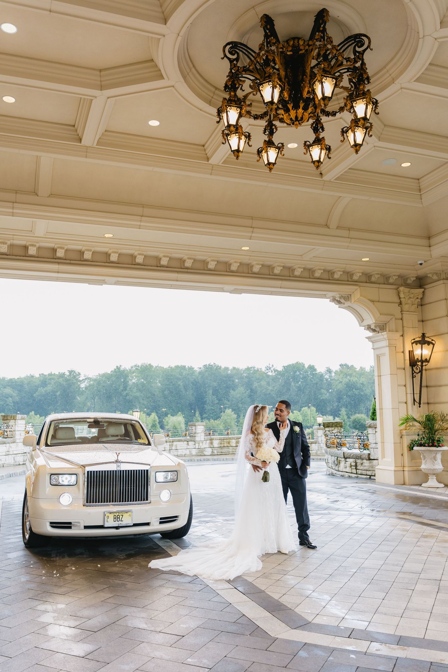 Bride and groom pose beside a white Rolls-Royce under an elegant chandelier at a luxury wedding venue.