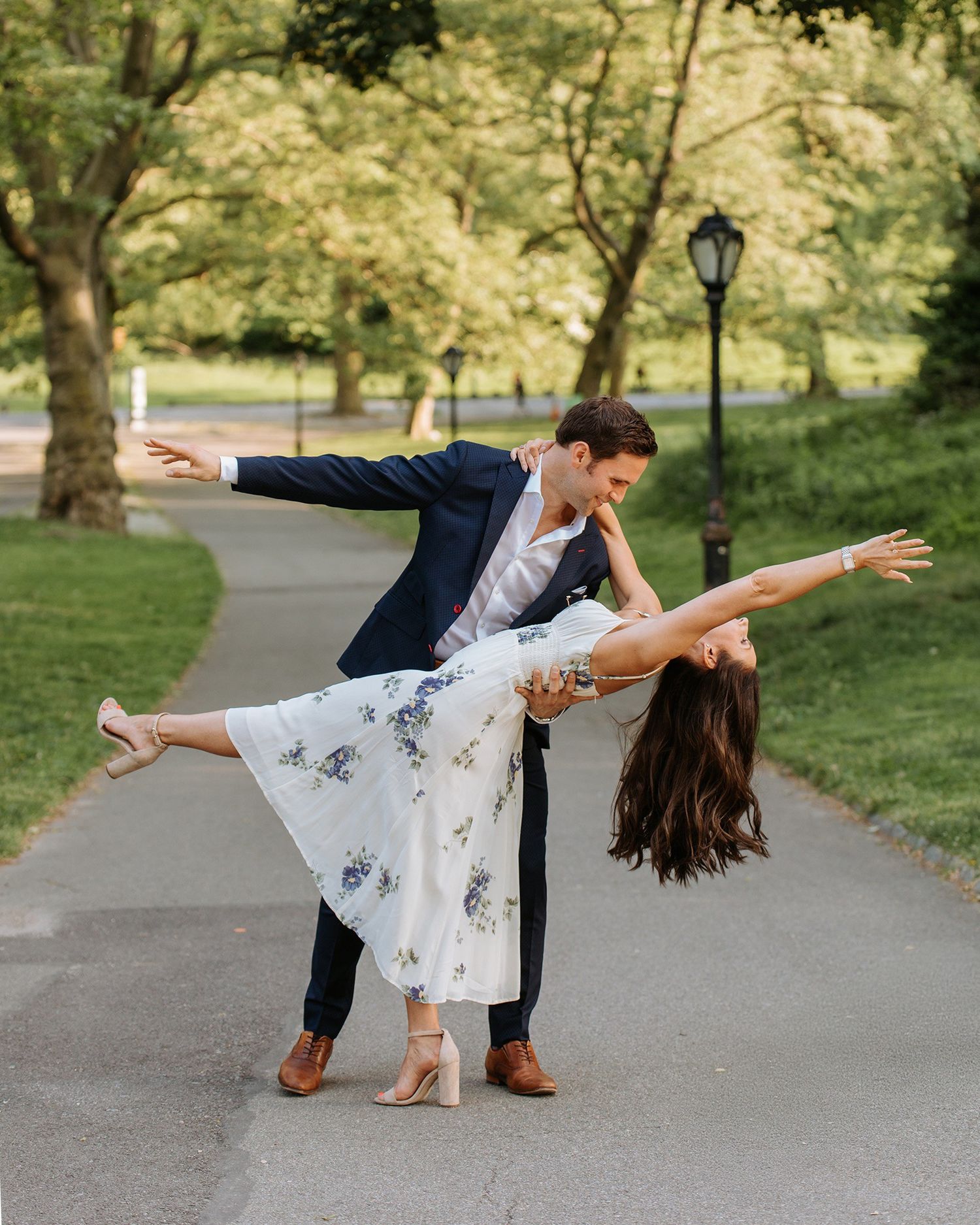 Couple performs romantic dance dip on a park pathway surrounded by green trees.