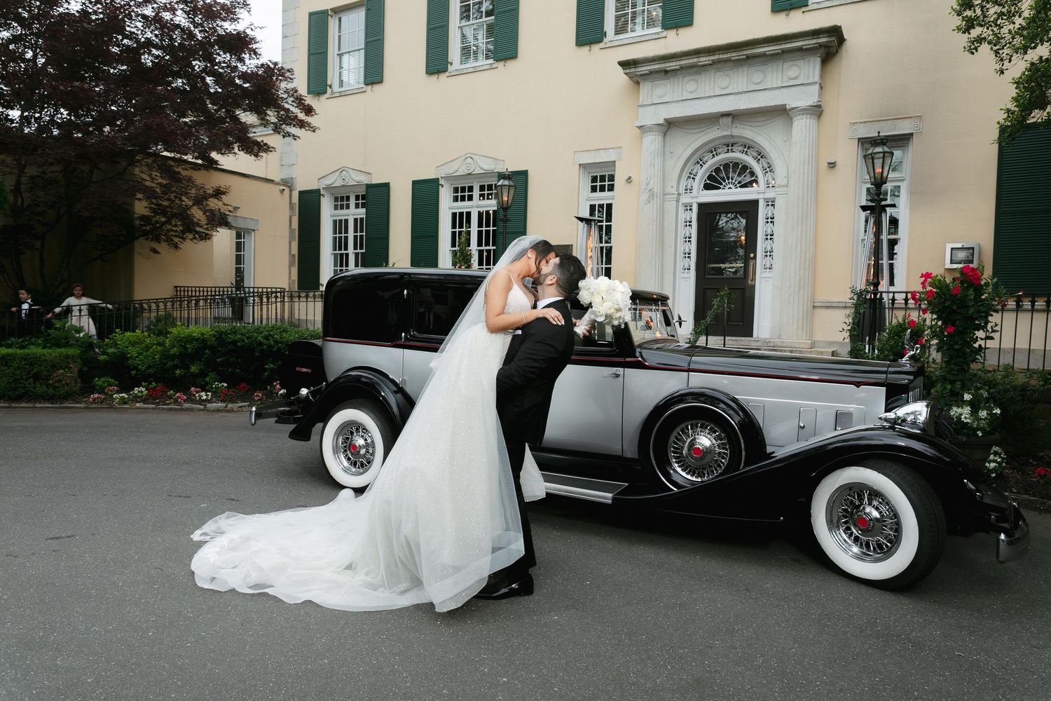 Classic vintage car parked outside an elegant mansion with white wedding dress flowing behind.