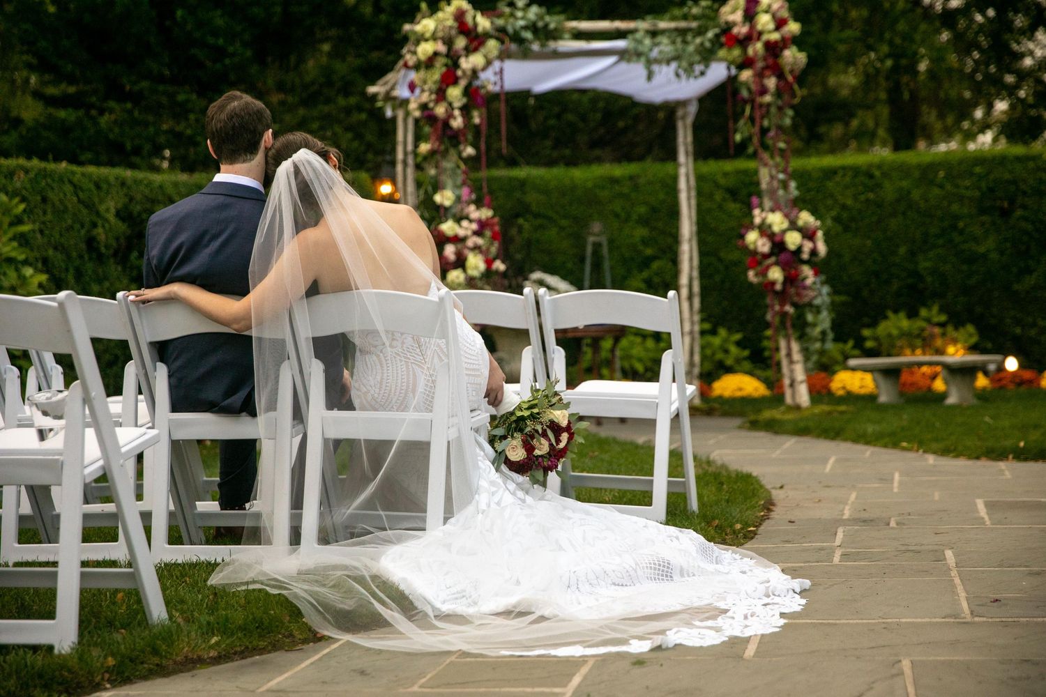 Wedding ceremony setup with white chairs and floral arch in an outdoor garden setting.