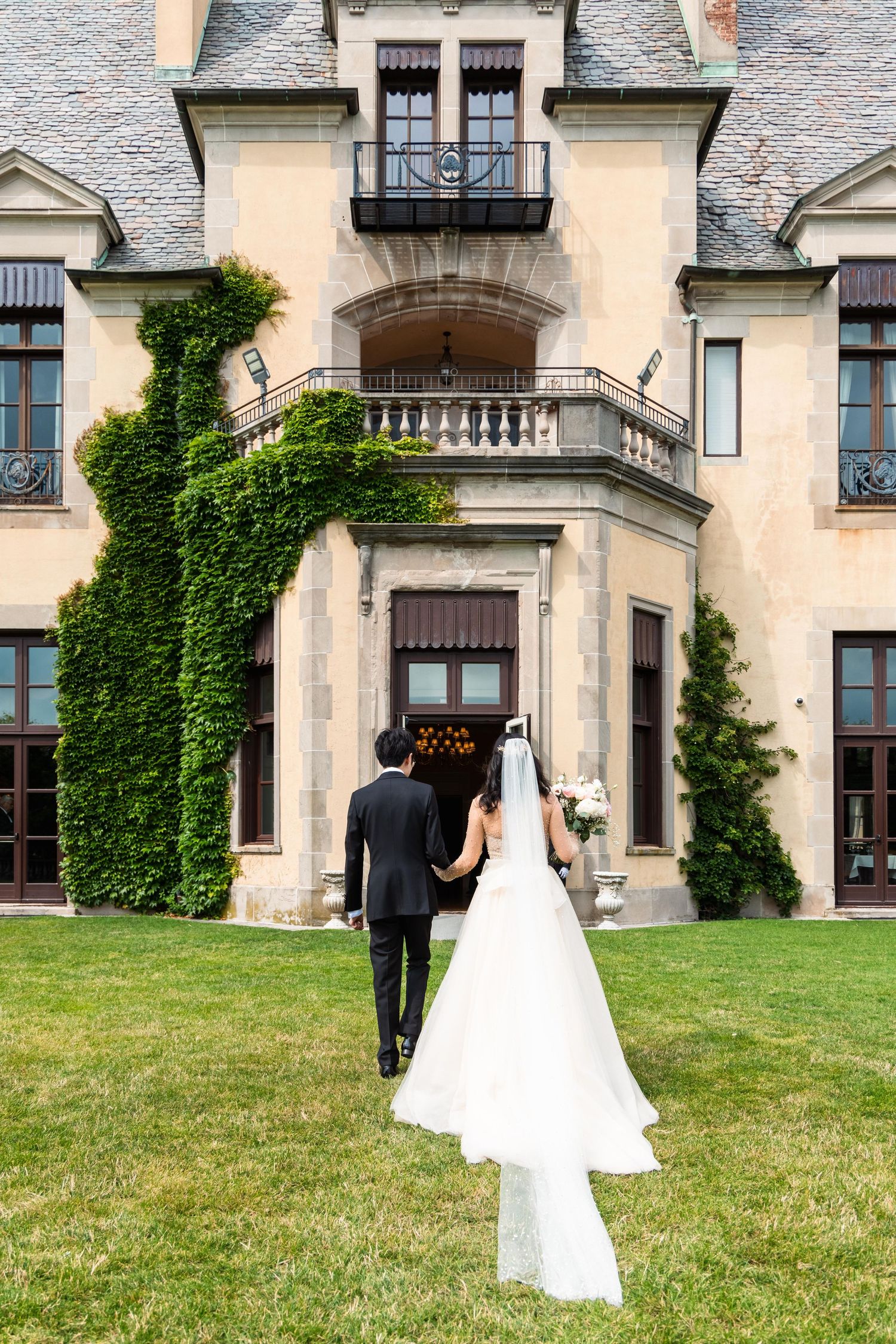 Wedding couple walking across manicured lawn of historic mansion with ivy-covered walls.