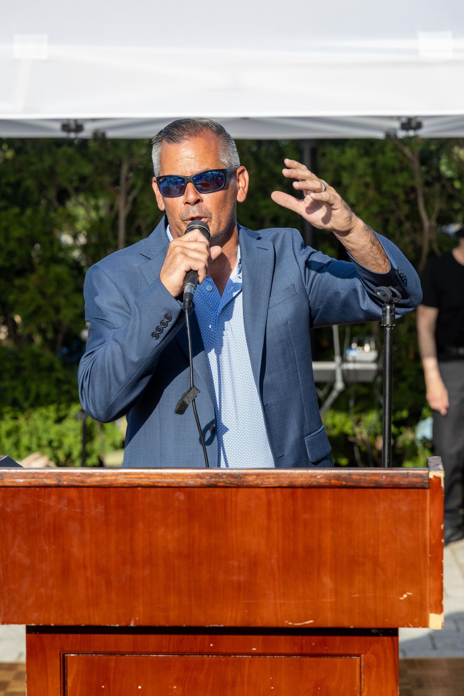Man in blue blazer and sunglasses speaks at podium with microphone at outdoor event.