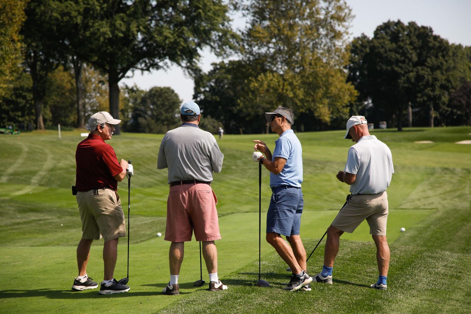 Four golfers stand together on a lush golf course fairway, holding clubs and discussing their game on a sunny day.