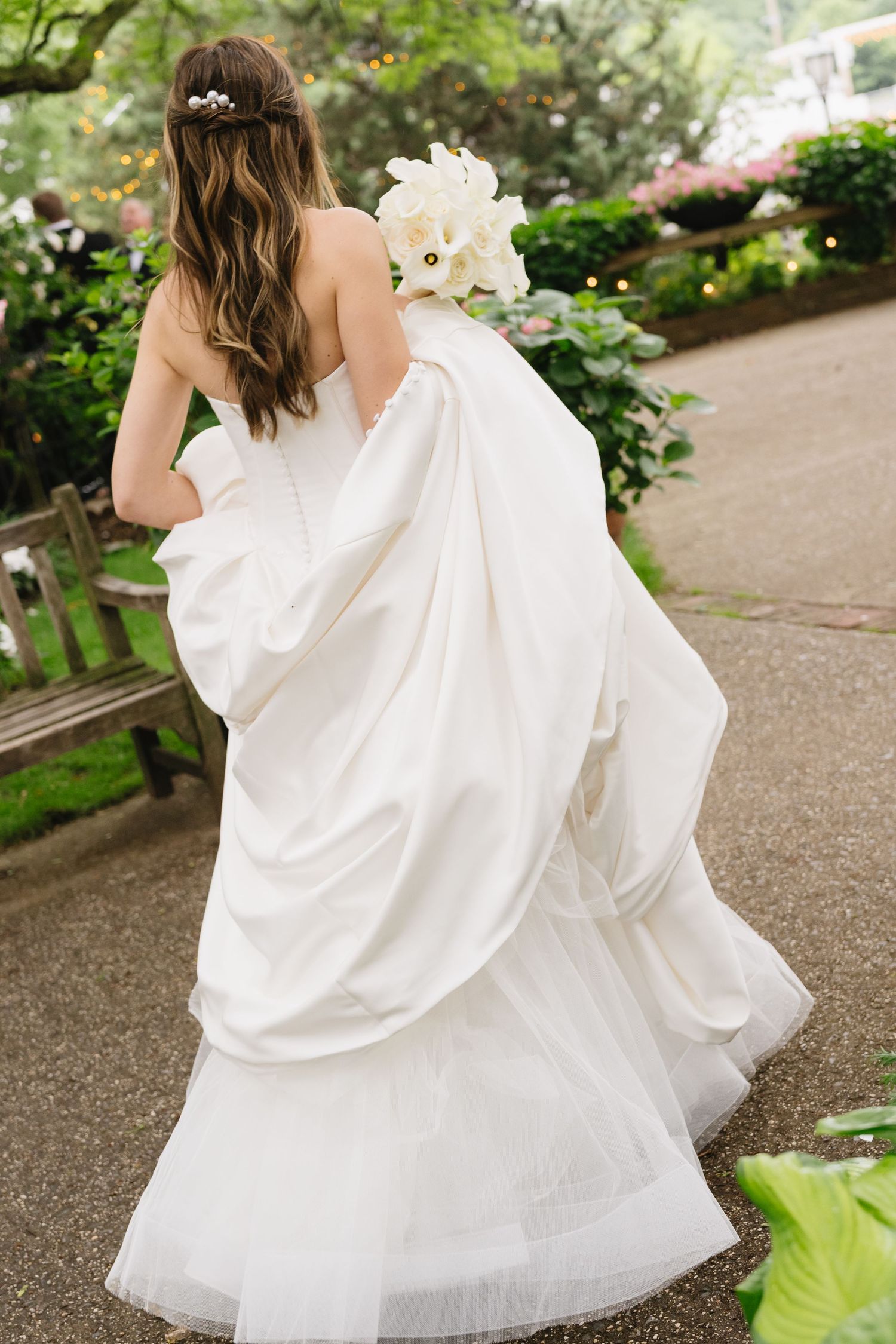 Bride in a flowing white strapless wedding gown holding a white bouquet walks through a lush garden pathway.