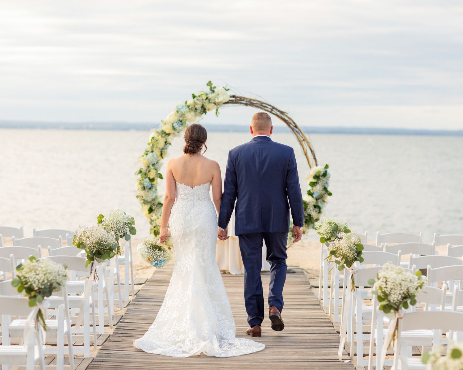 Couple walking down a scenic oceanfront pier decorated with white floral arrangements for a wedding ceremony.