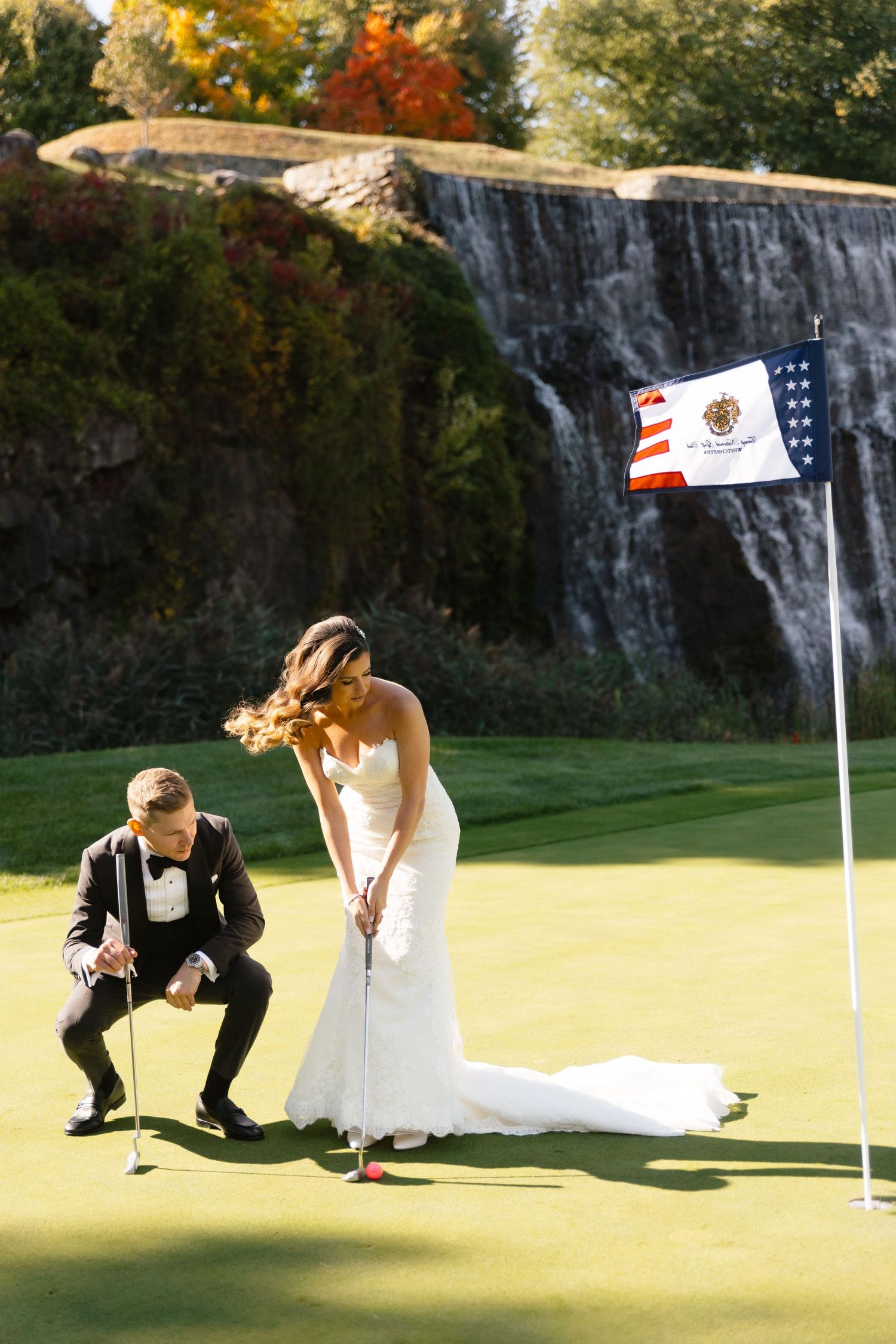 Wedding couple plays golf on putting green with waterfall and American flag in background.