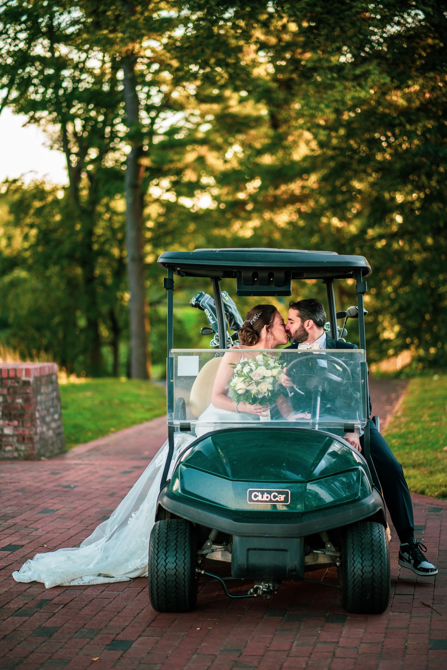 Wedding couple poses in a green golf cart on brick pathway surrounded by trees at sunset.