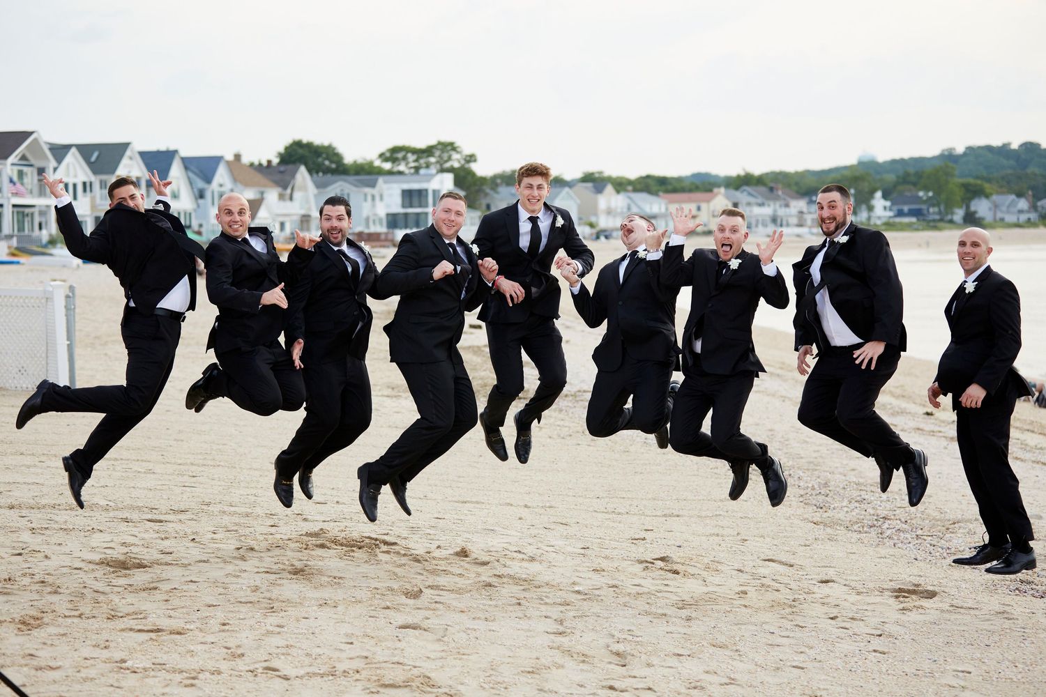 Large wedding party jumps in unison on a beach for a fun group photo.