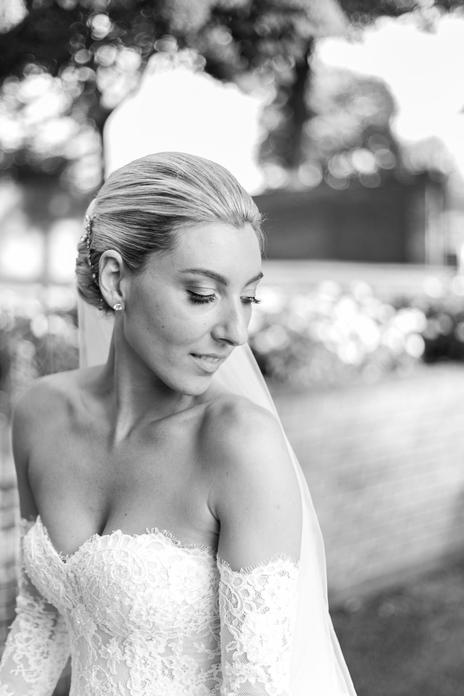 Elegant bride in lace off-shoulder wedding dress with flowing veil in black and white portrait.