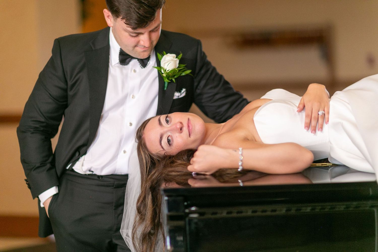Playful wedding couple poses on grand piano in formal attire.