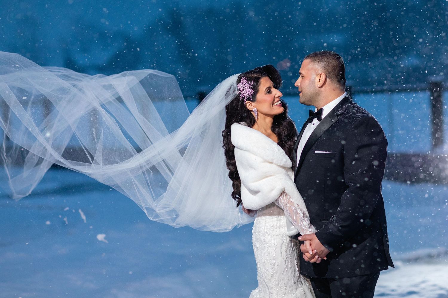 A winter wedding portrait with flowing veil and falling snow against a blue background.