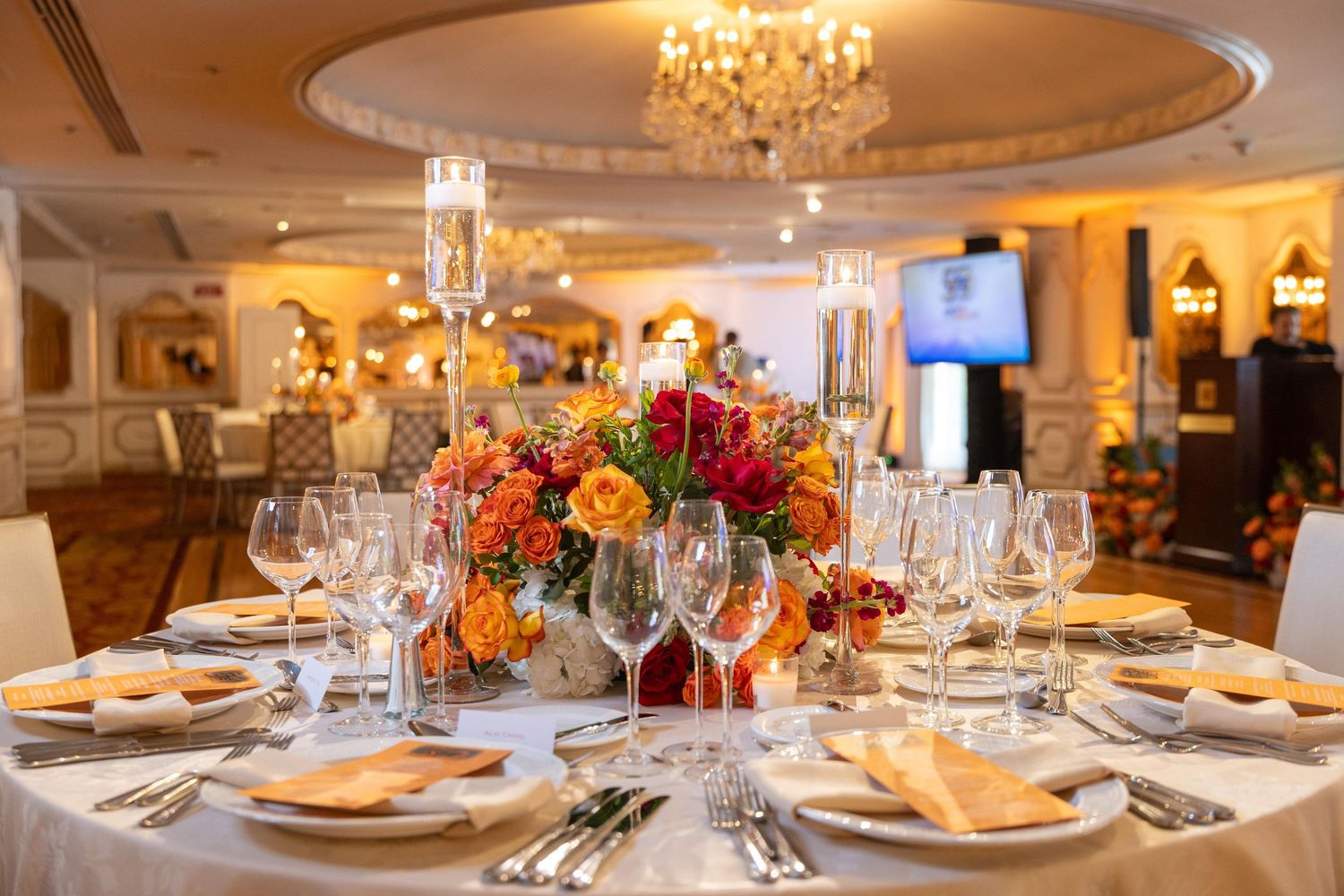 Elegant banquet table with orange and red floral centerpiece, crystal chandelier, and fine dining settings in a luxury ballroom.