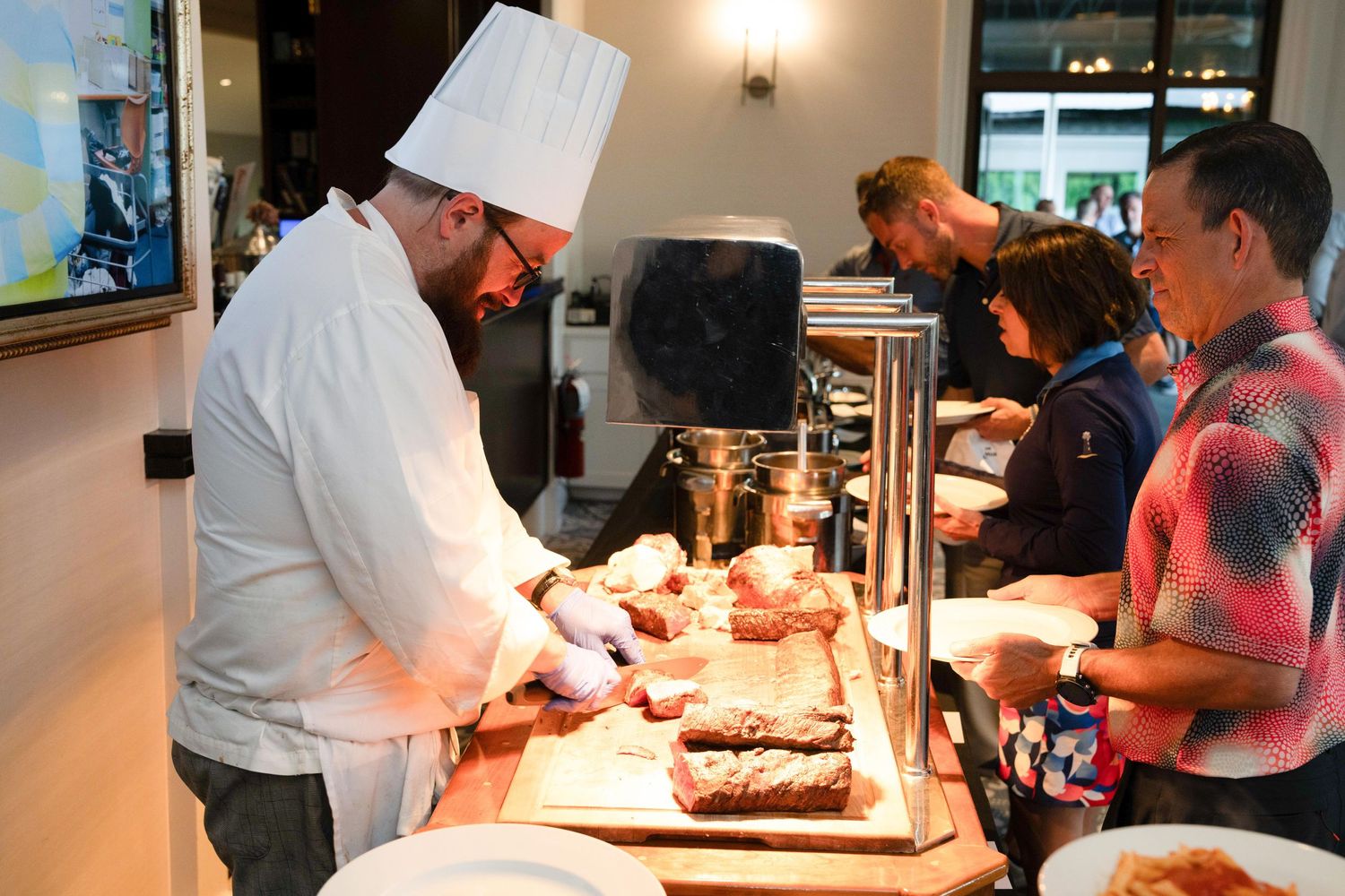 Chef in white uniform carves meat at a buffet station while guests wait in line at an event.