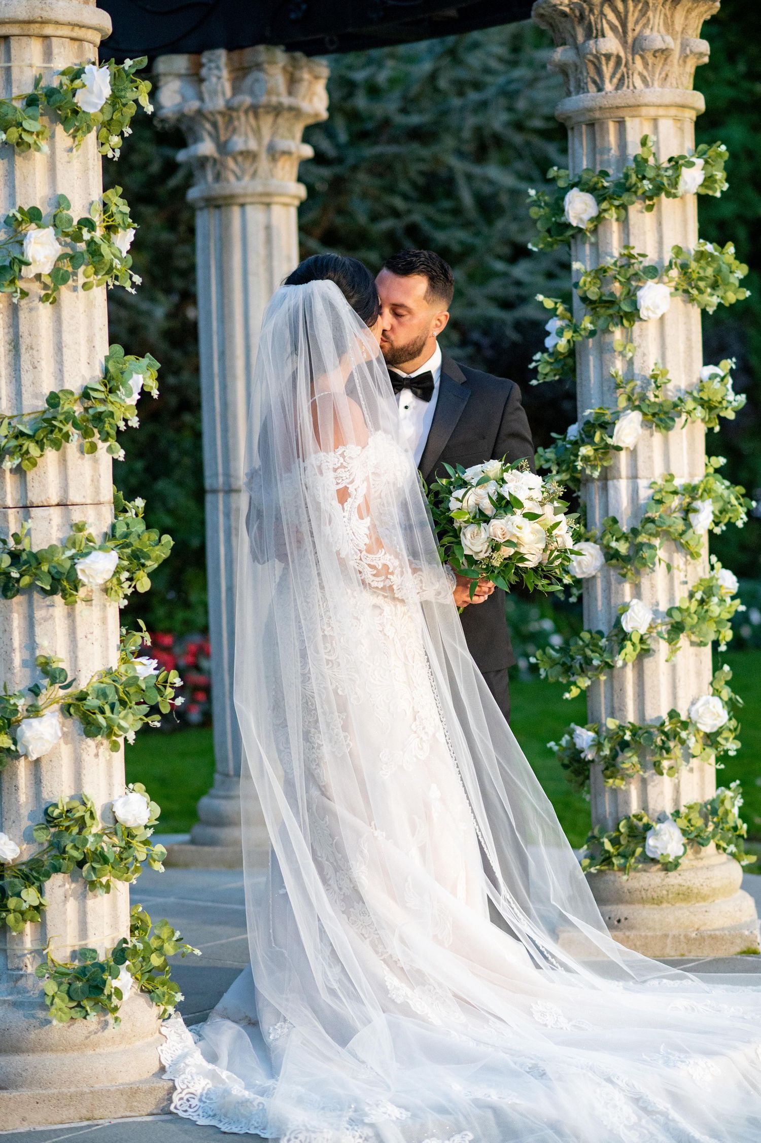 Elegant wedding portraits with flowing veil among decorated stone columns with floral garlands.