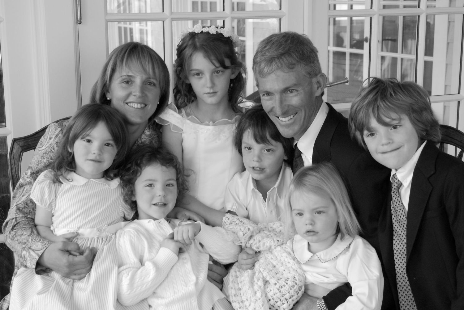 A group of people pose together in formal attire at an indoor gathering with white decor and windows visible.