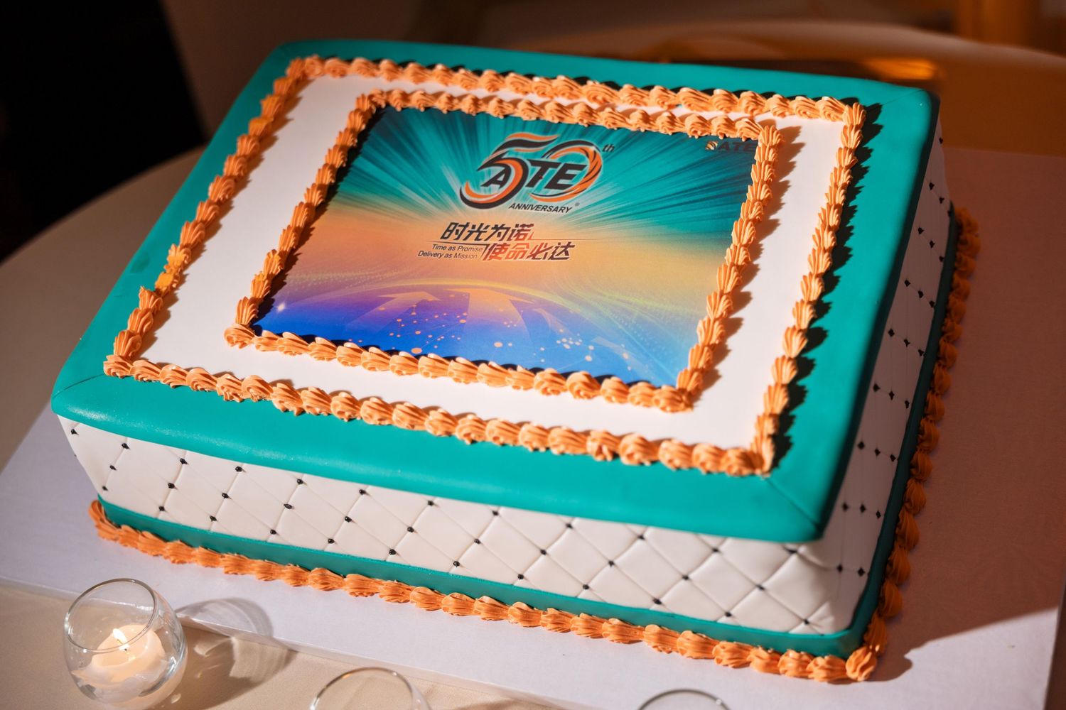 Elegant white and teal fondant celebration cake with gold piping and custom logo image topper on display.