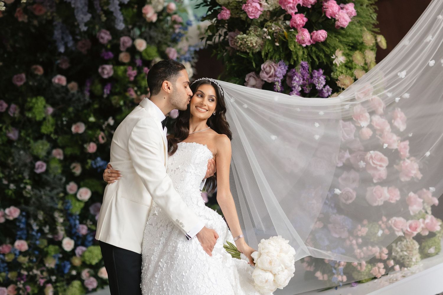 Wedding couple poses against floral arch with flowing veil and white bouquet.