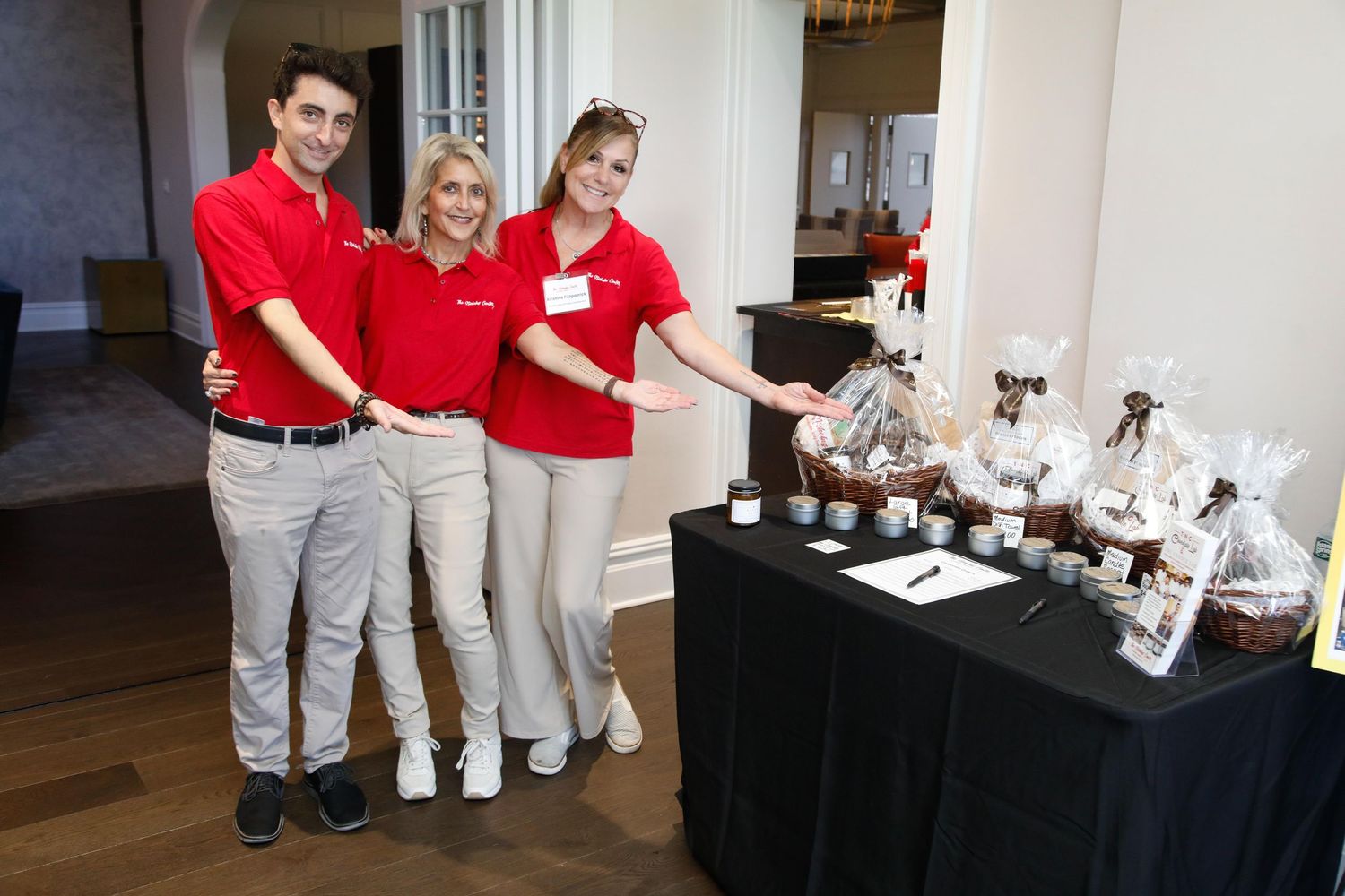 Three staff members in red shirts smile at a prize table with gift baskets at a charity event.