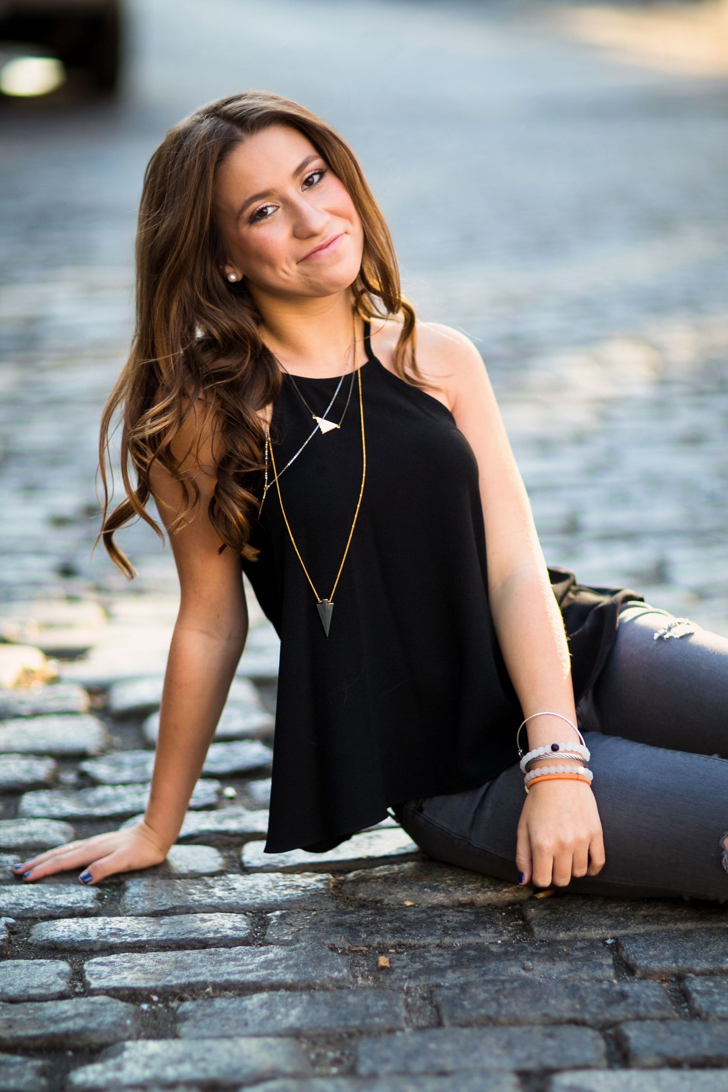 A person in a black sleeveless top and jeans sits on cobblestone pavement near water at sunset.