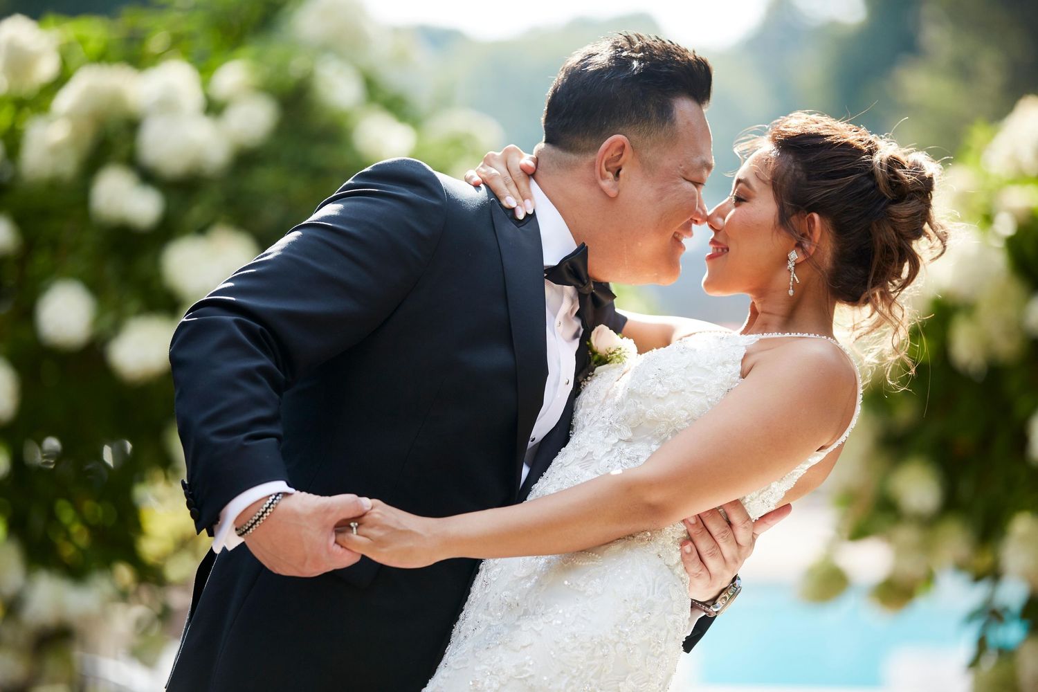 Romantic outdoor wedding portraits with couple embracing among white flowers and greenery.