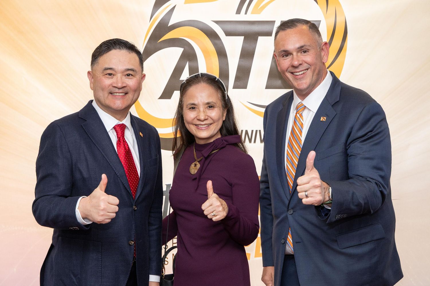Three smiling professionals give thumbs up in front of a 50th anniversary logo backdrop at a formal event.