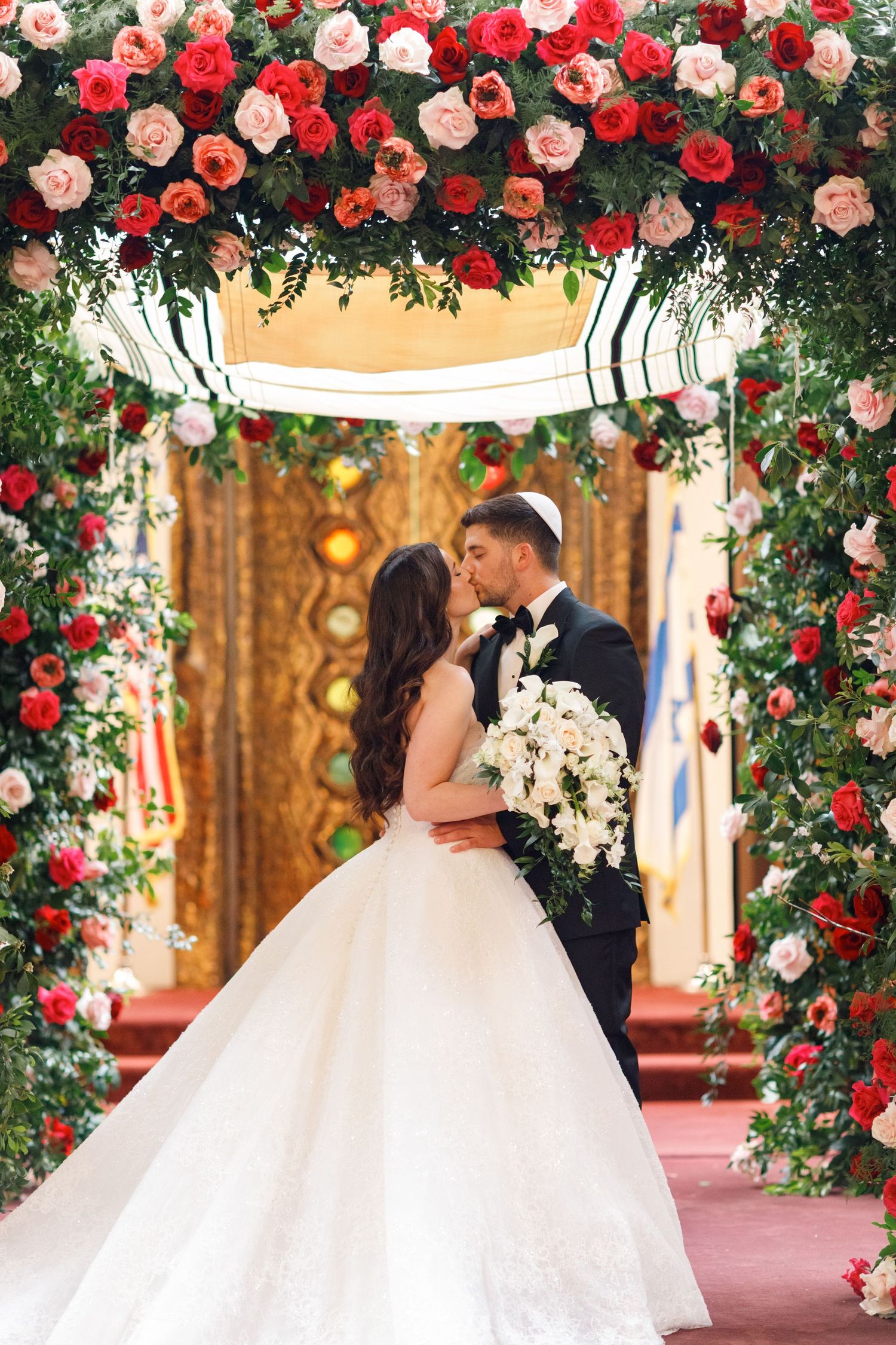 Bride and groom kissing under a floral chuppah adorned with red and pink roses at a Jewish wedding ceremony.