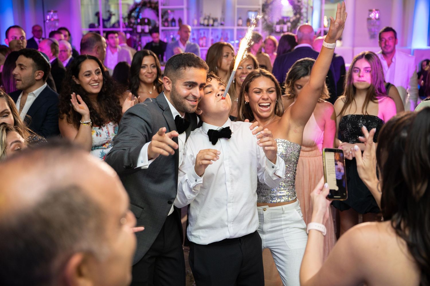 Joyful wedding reception dance floor with guests celebrating, man in tuxedo and woman in silver dress dancing.