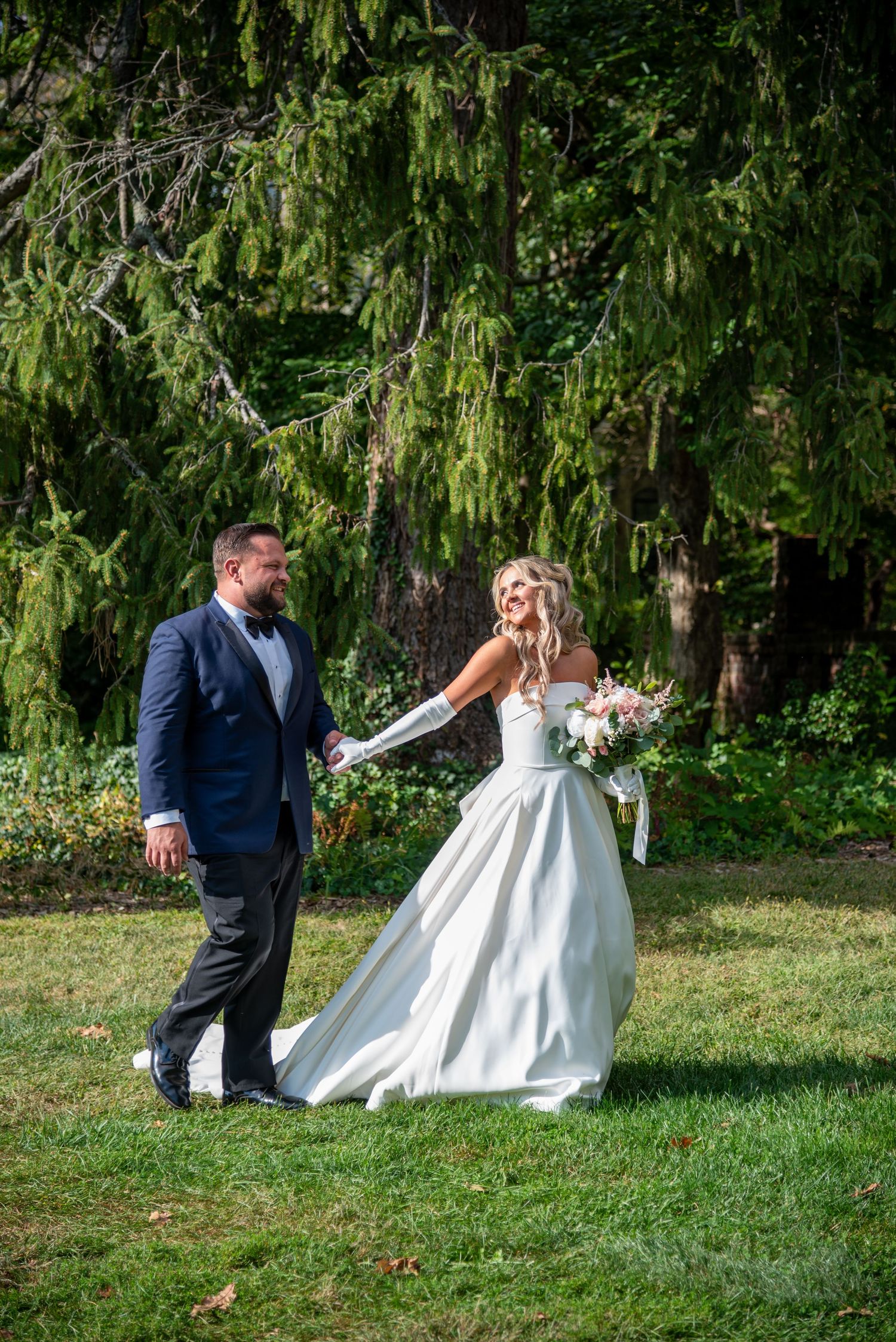 Playful wedding couple dancing on grass beneath towering willow trees.