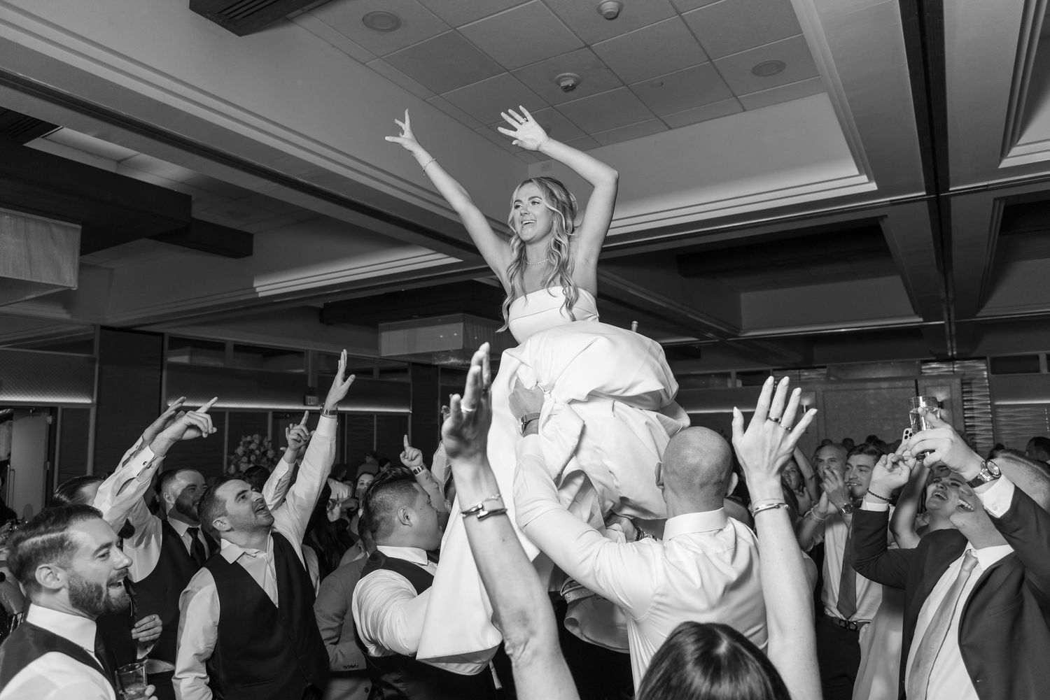 Bride being lifted up by wedding guests during lively dance floor celebration.