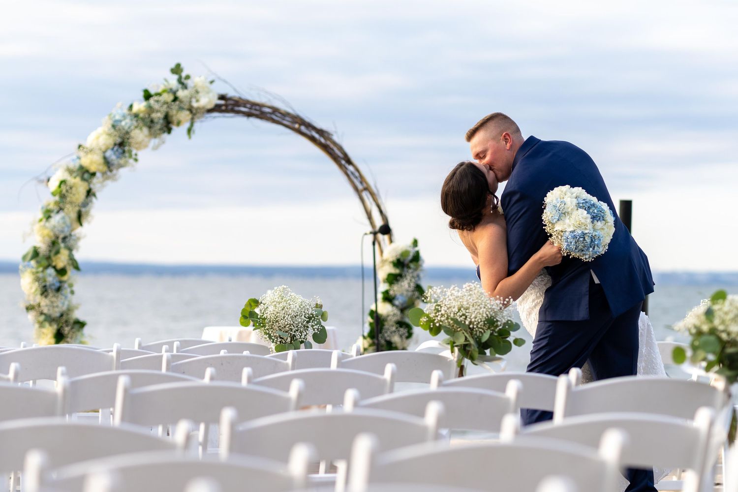 White chairs set up for an outdoor wedding ceremony with floral arch decorations.