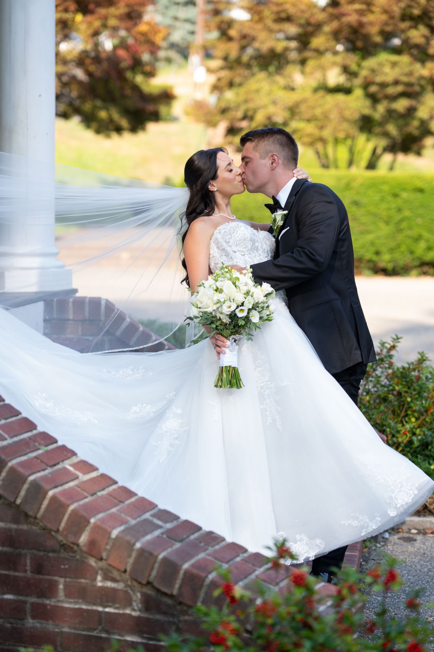 A couple shares romantic moments near white columns and brick walls with flowers.
