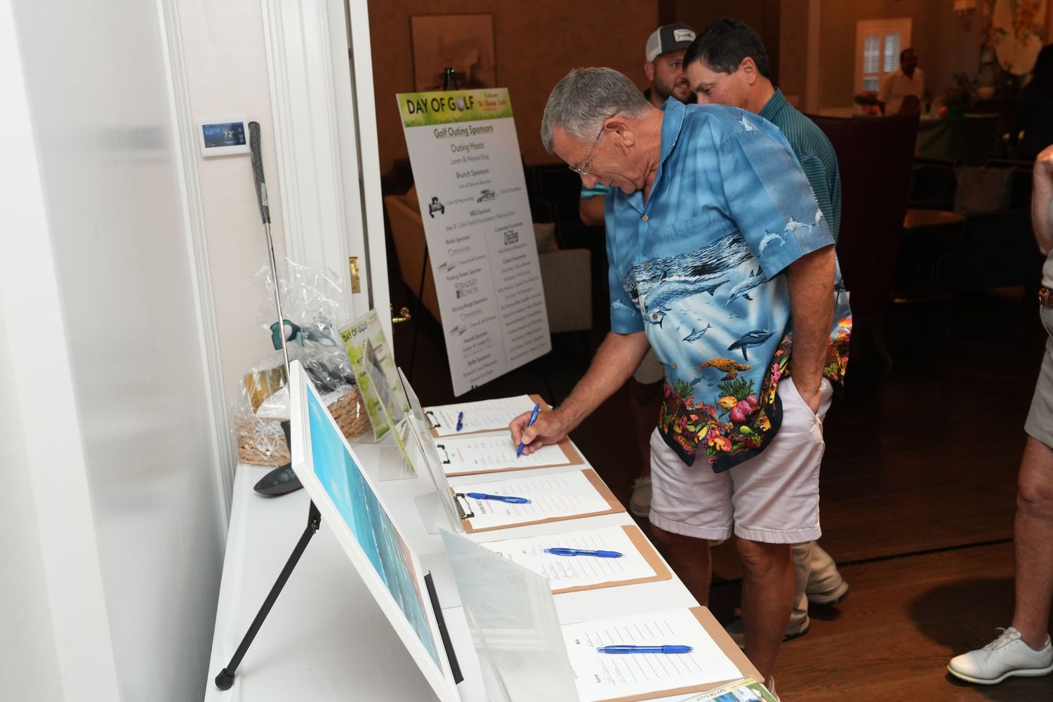 Man in blue floral shirt signs documents at a registration table during an outdoor charity golf event.