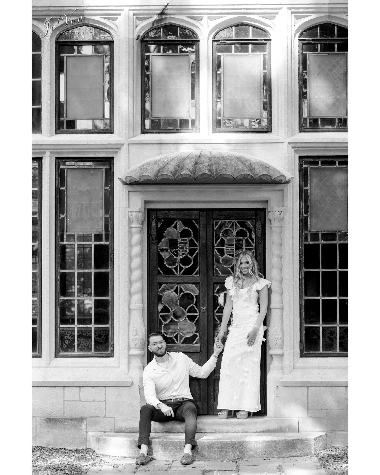 Black and white portrait of couple posed in front of historic building with ornate windows.