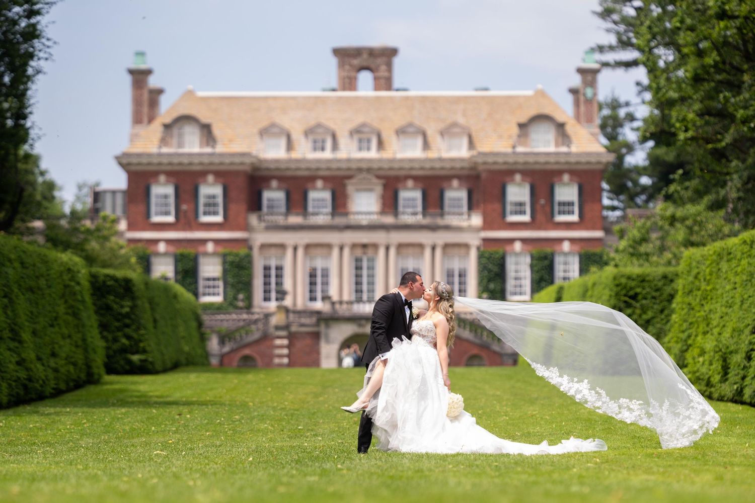 Elegant wedding couple poses in front of a grand mansion with flowing veil caught in the breeze.