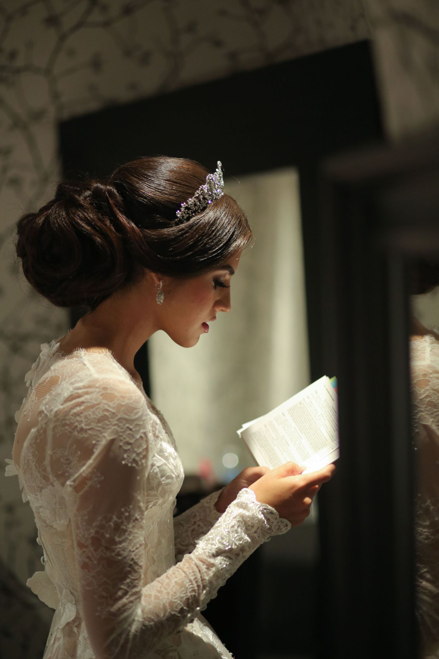 Bride in lace gown and tiara reads from illuminated book in elegant setting.